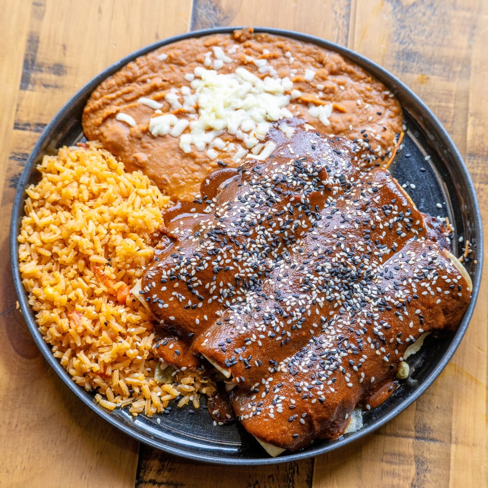 Three enchiladas covered in mole sauce and sesame seeds, with rice and refried beans on a black plate.