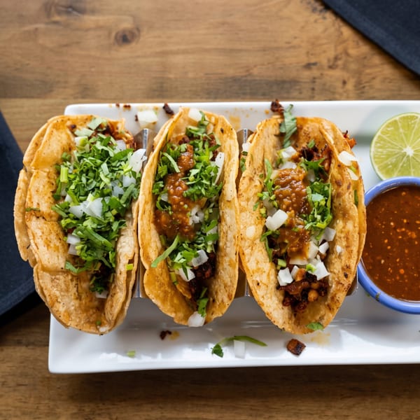Three street tacos with meat, salsa, onions, and cilantro, served with a lime wedge and red salsa.