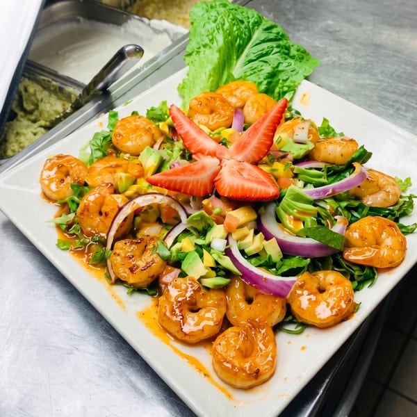 Shrimp salad with avocado, red onion, and strawberries on a white plate in a commercial kitchen.