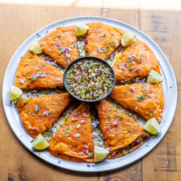 A round platter of birria quesadillas, garnished with red onion, cilantro, lime wedges, and a central dipping sauce.
