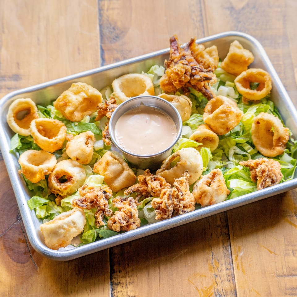 Fried calamari and onion rings on a bed of lettuce with a dipping sauce in a metal tray.