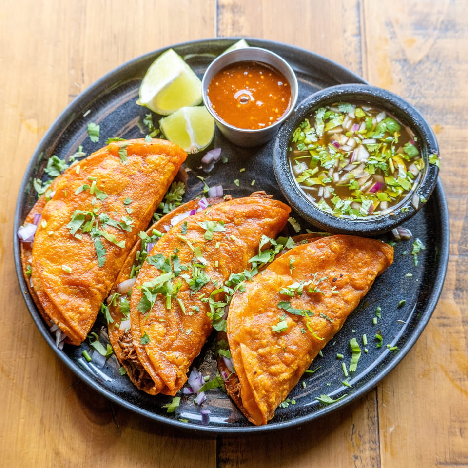 Three crispy birria tacos with cilantro, red onion, lime wedges, and two dipping sauces on a black plate.