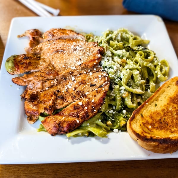 Grilled chicken with green pasta and a slice of toasted bread on a white plate.
