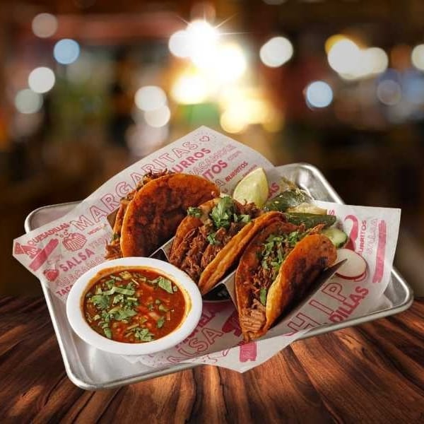 A metal tray with four birria tacos, a side of consommé, lime, and pickled vegetables.