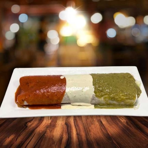 Burrito covered in red, white, and green sauces, resembling the Mexican flag, on a white plate.