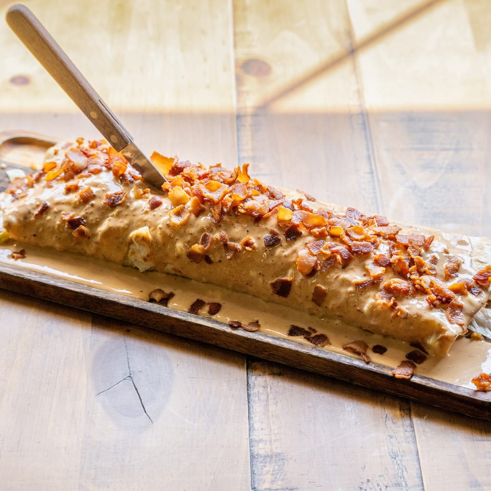 A large burrito covered in creamy sauce and bacon bits, with a knife stuck in it, on a wooden tray.