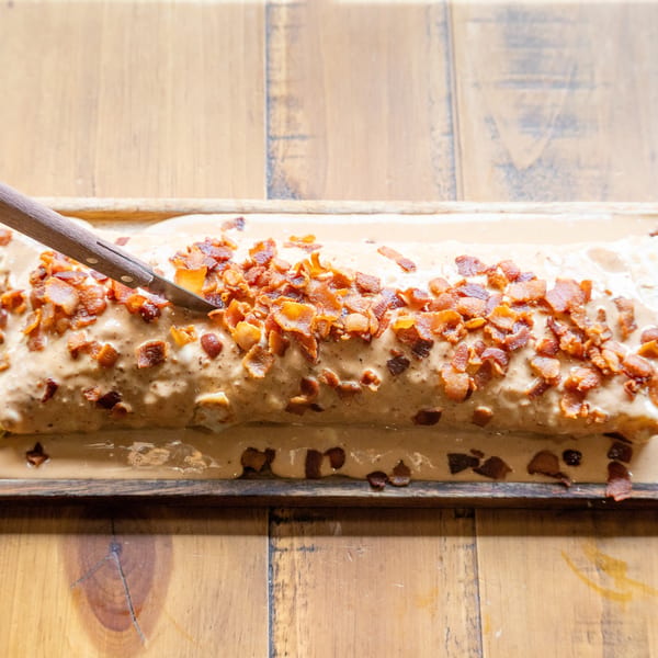 A long burrito covered in creamy sauce and crispy bacon bits, with a knife stuck in it.