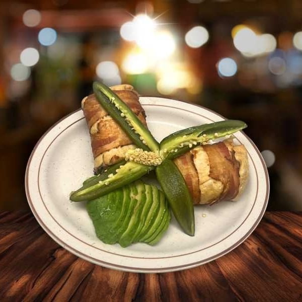 Bacon-wrapped chicken with sliced jalapeños and avocado on a white plate, on a wooden table.