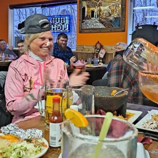 A woman in a pink hoodie laughs at a table with food and drinks at a lively restaurant.