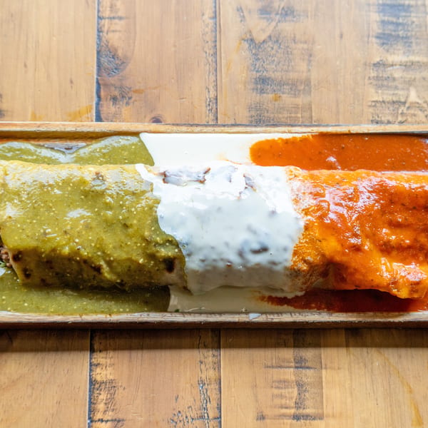 A large burrito covered in green salsa, white sour cream, and red salsa on a wooden tray.
