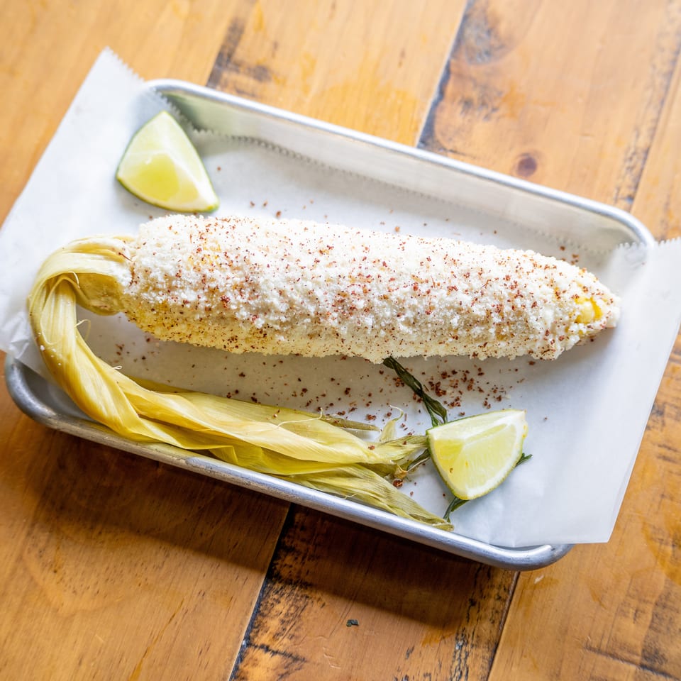 Elote, Mexican street corn, on a metal tray with parchment paper and lime wedges.